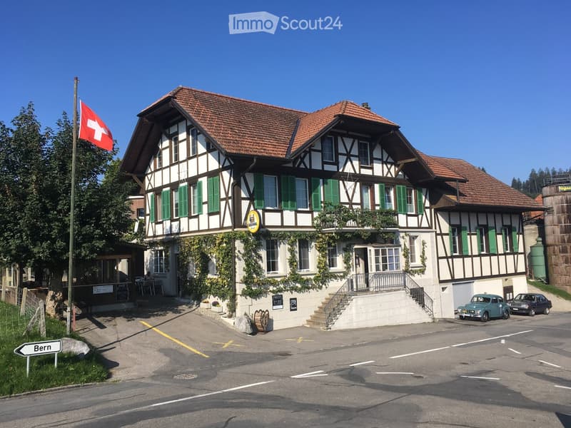 Traditional Swiss building with a red-tiled roof, green shutters, and a balcony adorned with plants. A Swiss flag is displayed on a pole, and a sign reading "Bern" is visible on the left side.