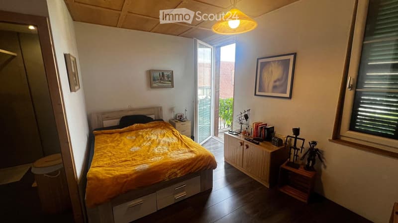 Cozy bedroom with a wooden bed, yellow bedspread, nightstand, cabinet, and artwork on the walls. The room features wooden flooring and a hanging light fixture.