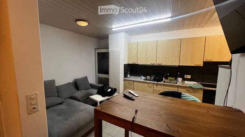 A modern, open-plan living and kitchen area with a grey sectional sofa, a wooden coffee table, and a kitchen with light wooden cabinets and a black tiled backsplash.