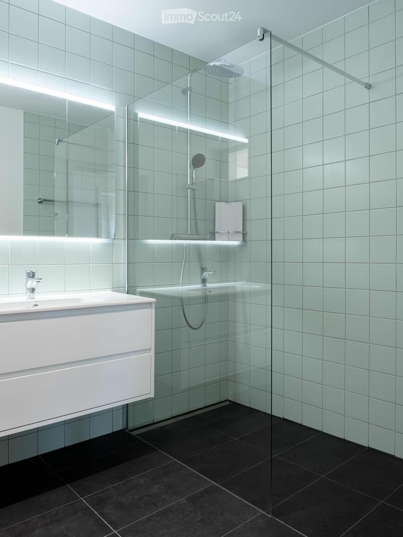 Modern, minimalist bathroom with green-tiled walls, black floor tiles, white vanity, glass shower enclosure, and overhead LED lighting