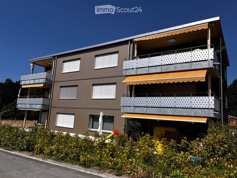 Three-story building with brown facade, windows, and balconies with yellow awnings. Shrubbery and plants in front.