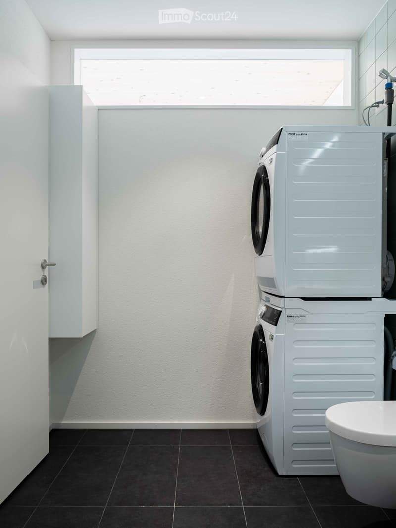 Modern utility room with white tiled flooring, two stacked washing machines, a white toilet, and a window.