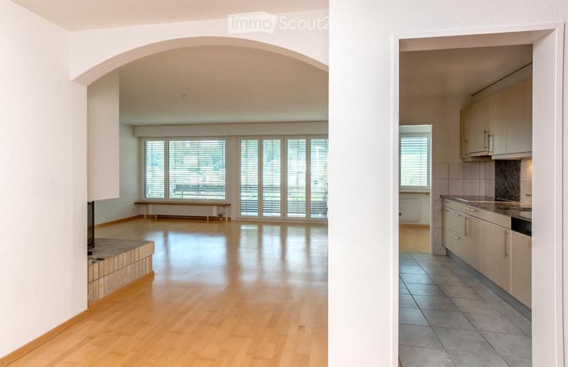 The image shows an unfurnished living room and kitchen area. The living room has wooden flooring, large windows with blinds, and a fireplace. The kitchen features light-colored cabinets, a tiled backsplash, and a countertop.