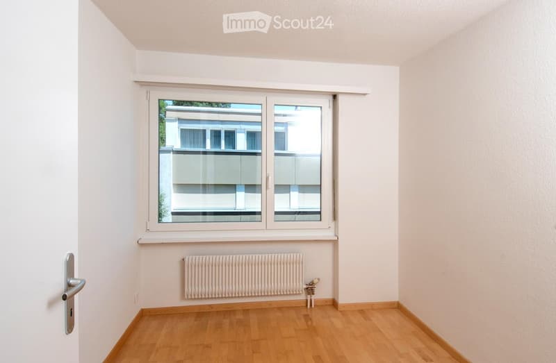 Empty room with light-colored walls, wooden flooring, a window with white frames, and a radiator below the window.