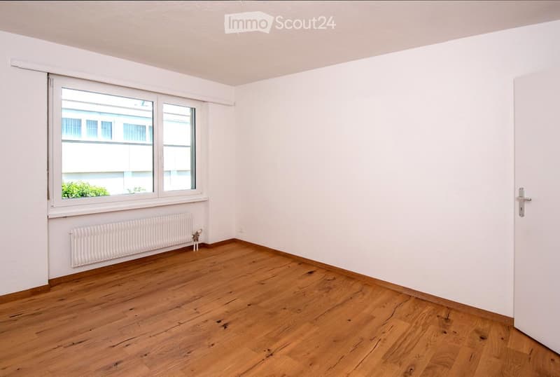 Empty room with wooden flooring, white walls, a window with white frame, and a radiator below the window.
