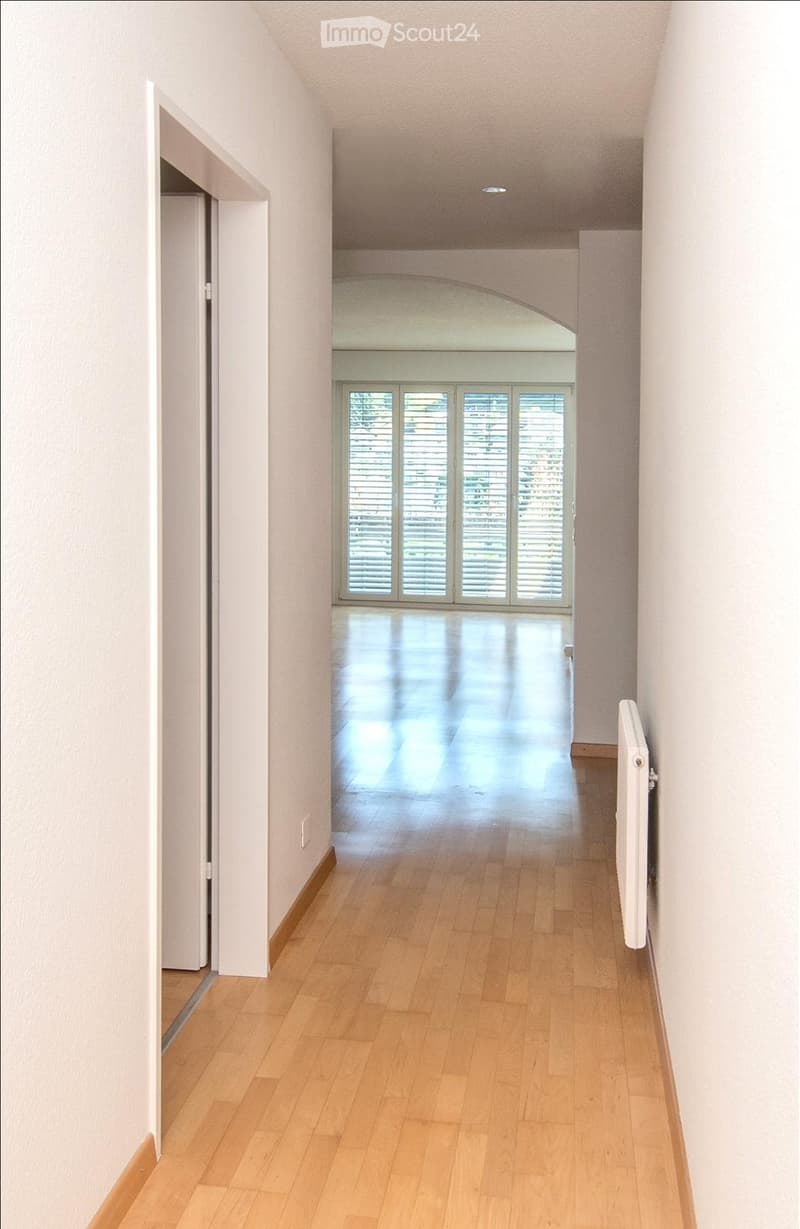 Empty hallway with light wooden flooring, white walls, and a radiator on the right side. An open doorway leads to a room with large windows and a shiny floor.