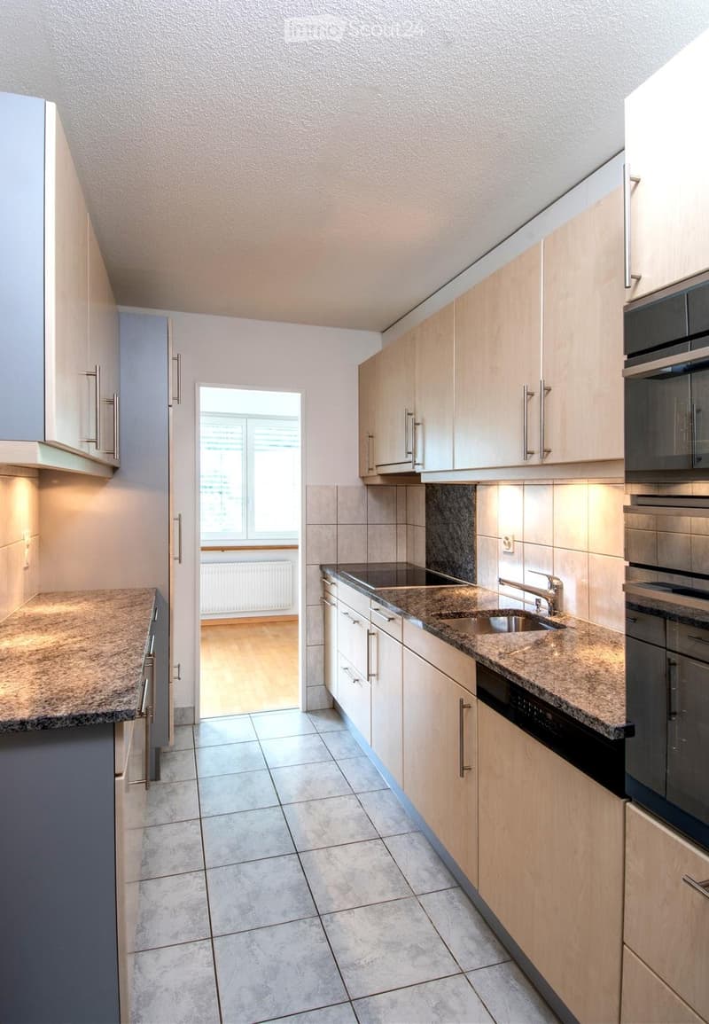 Modern kitchen with light wooden cabinets, granite countertops, and stainless steel appliances including a sink, faucet, oven, and dishwasher. The space features tiled flooring and a window leading to another room.