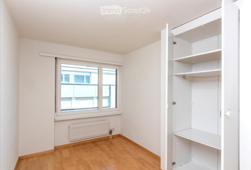 Empty room with wooden flooring, white walls, a window with a view, a radiator, and an open built-in closet with shelves and a hanging rod.