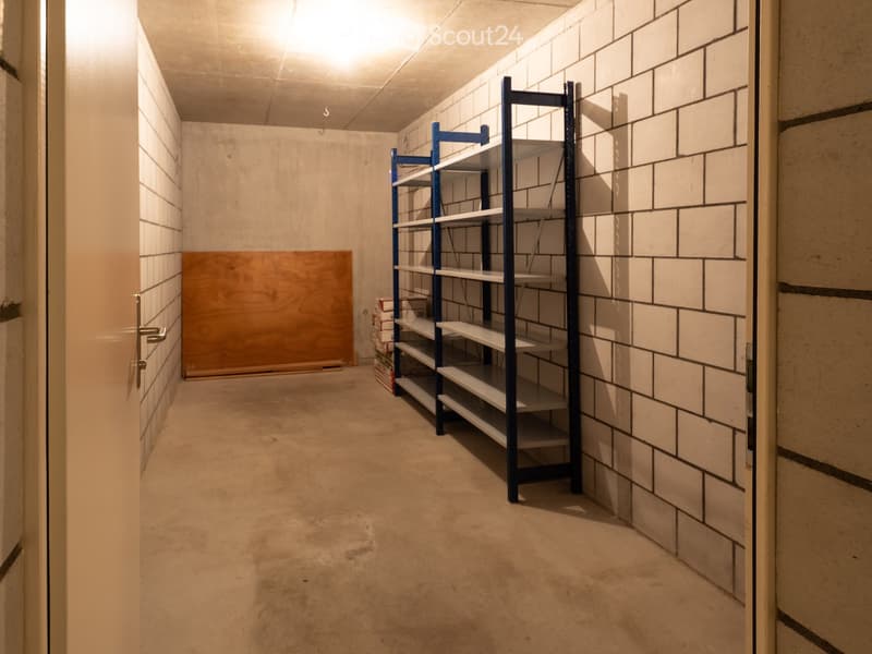 Empty storage room with concrete floor, white tiled walls, and a metal shelving unit. A wooden board leans against the wall, and a stack of boxes is placed near the shelving unit.