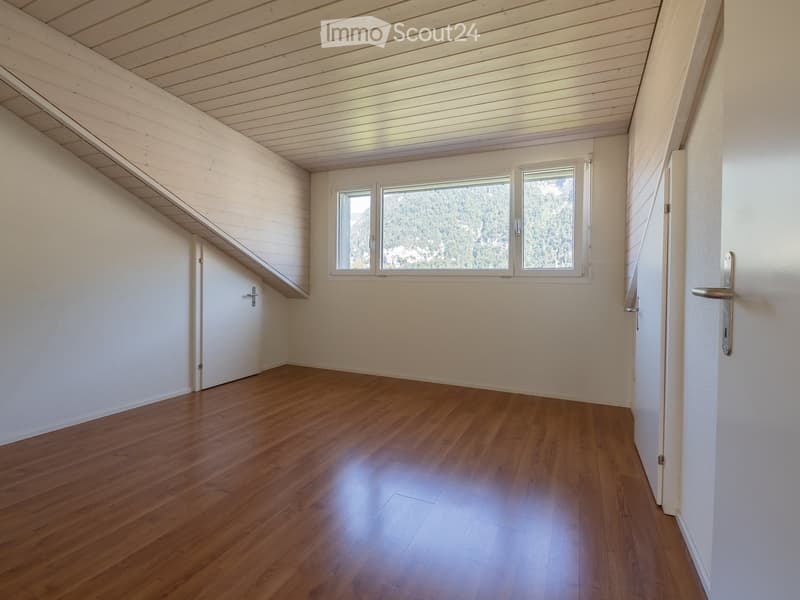 Empty room with wooden flooring, white walls, and large windows offering a scenic view. The room features a slanted wooden ceiling and a small closet with a door.