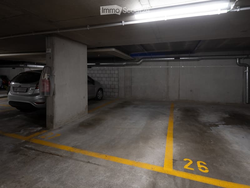 Underground parking area with marked parking spaces, two cars parked, concrete pillars, and fluorescent lighting.