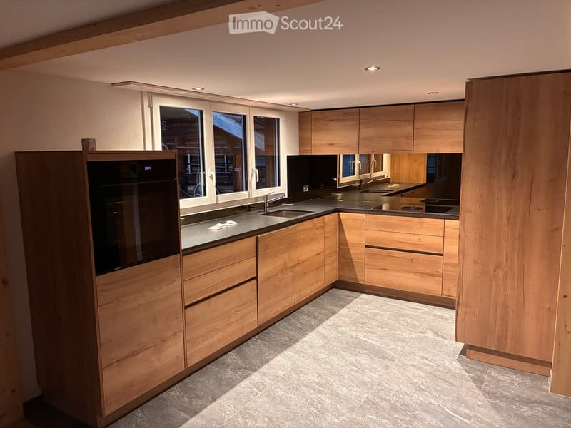 Modern kitchen with light wooden cabinets, stainless steel sink, and built-in oven. Large windows provide natural light.