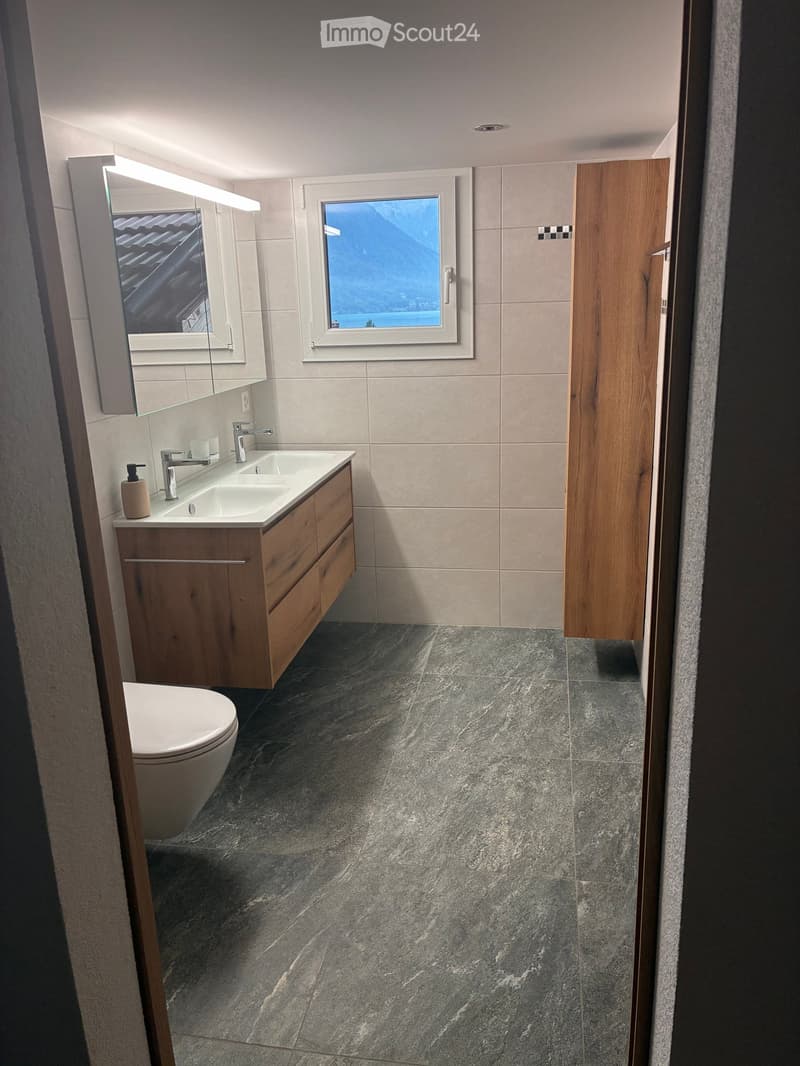 Modern bathroom with a double sink, wooden cabinet, mirror, and a toilet. Features a large window with a scenic view and grey marble flooring.