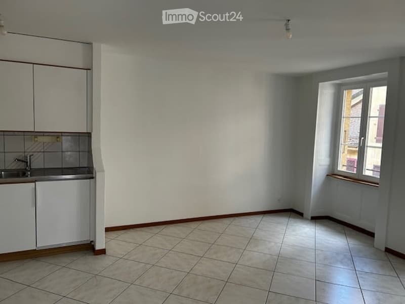 Empty room with tiled floor, white walls, and a small kitchen area with cabinets and sink.