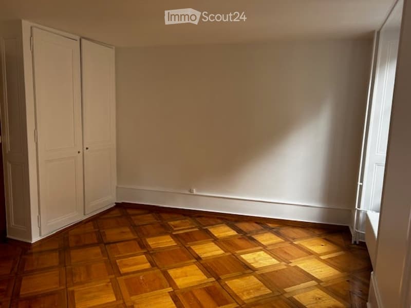 Empty room with wooden parquet flooring and white walls, featuring a built-in closet on one side and a window allowing natural light to enter.
