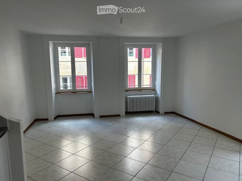 Empty room with tiled floor, white walls, two windows with red shutters, and a radiator under one window.