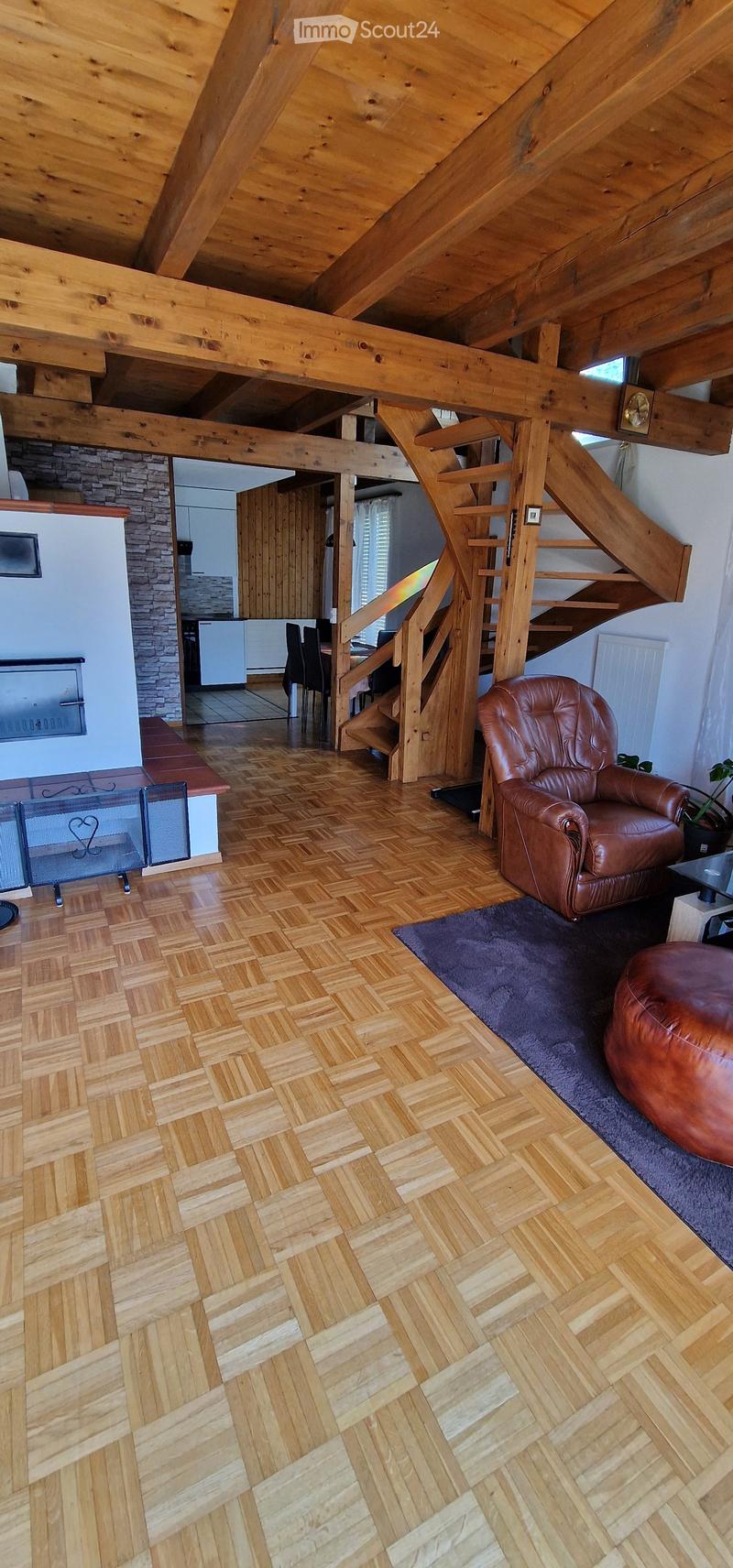 Living room with wooden herringbone flooring, exposed wooden beams, leather sofa, ottoman, dining area with chairs and table, fireplace, and staircase leading to another level.