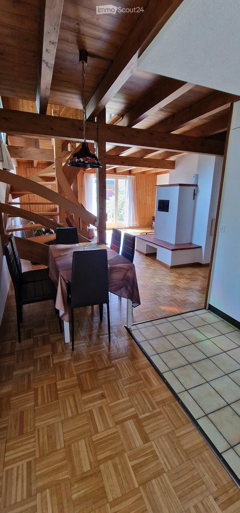 Dining area with wooden herringbone flooring, a dining table with chairs, a bench, and a refrigerator in the background. The ceiling features exposed wooden beams and a hanging light fixture.