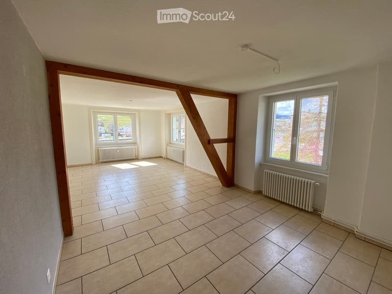 Empty room with tiled floor, white walls, and large windows. Radiators are visible along the walls.
