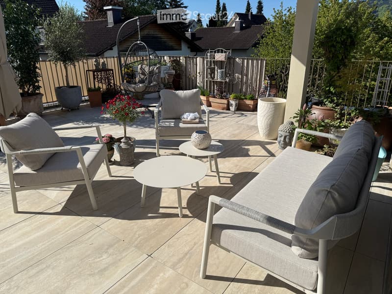 Well-furnished balcony with a sofa, chairs, tables, and decorative vases, surrounded by potted plants and flowers.