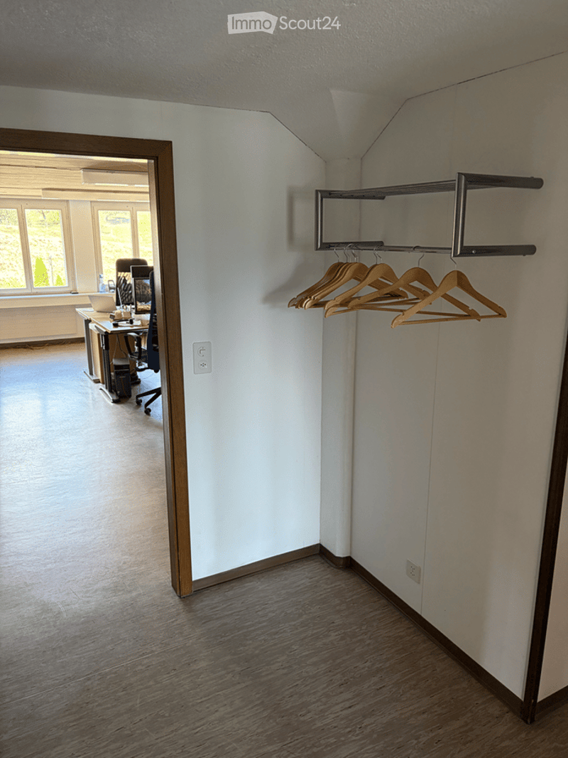 Empty room with light-colored walls, wooden flooring, and a coat rack with hangers. A doorway leads to another room with a desk and chairs.