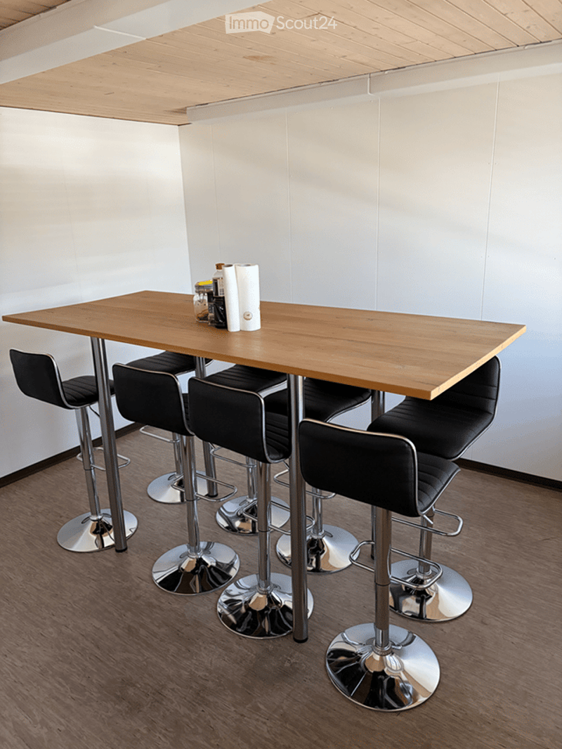 A modern, minimalist room with a wooden table and four black bar stools, featuring a clean, white wall background and a polished wooden floor.