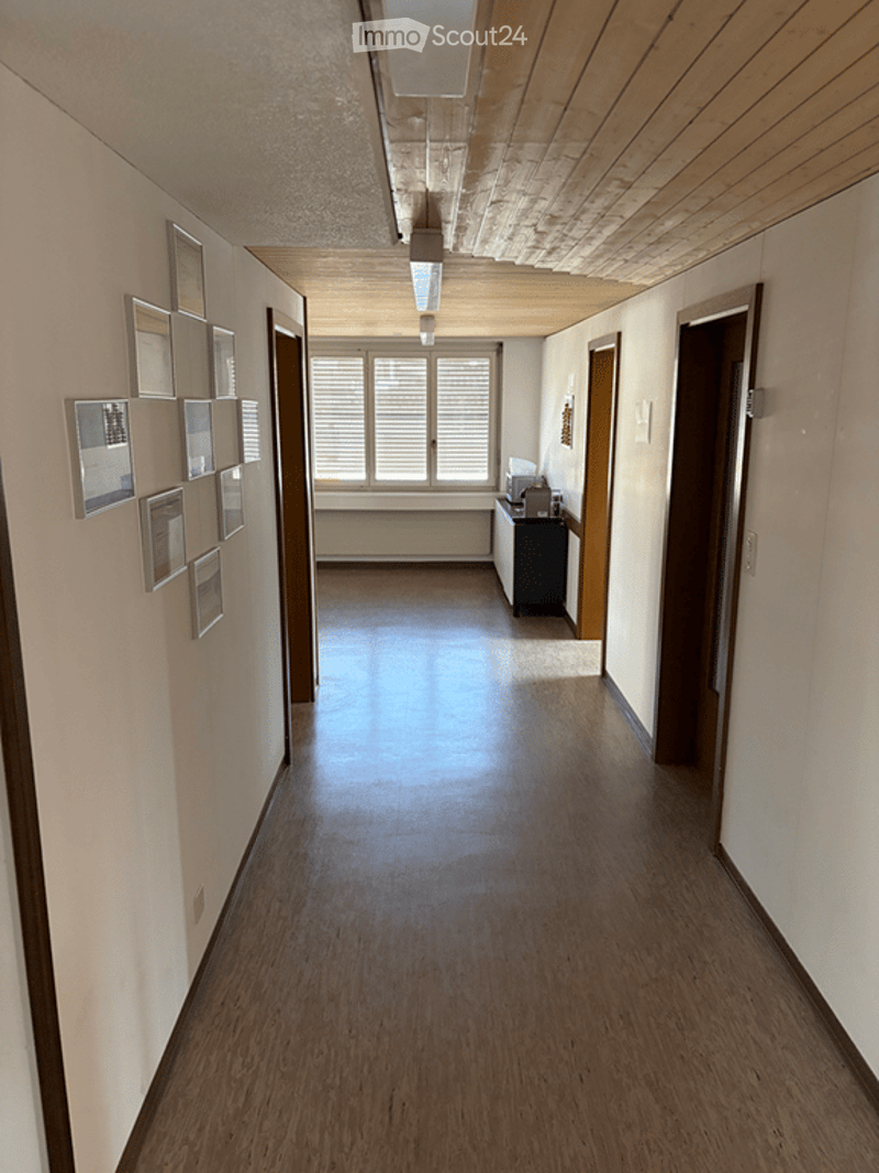 Empty hallway with wooden flooring, white walls, and several doors. A small cabinet and a window are visible at the end of the hallway.