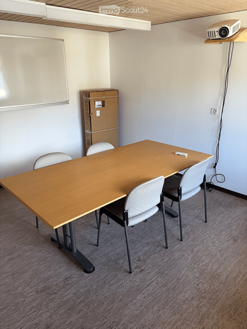 A modern meeting room with a large wooden table, several chairs, and a projector mounted on the wall. The room has a clean, minimalist design with a whiteboard and a cardboard box in the corner.