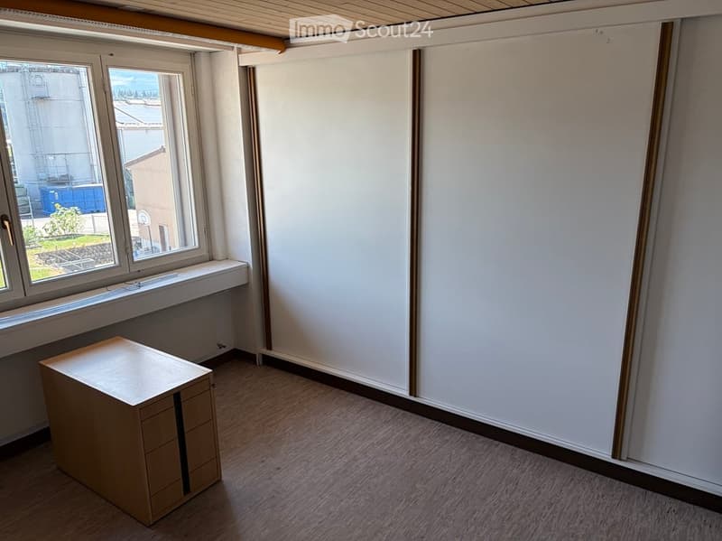 Empty room with wooden flooring, white walls, and a large window with a view of the outside. A small wooden cabinet is placed near the window.