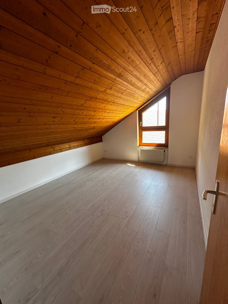 Empty attic room with wooden slanted ceiling, light wooden flooring, and a window with a radiator below it.