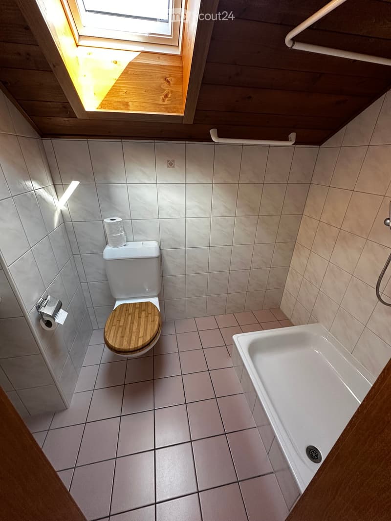 Modern bathroom with white tiled walls and floor, a toilet with a wooden seat, a bathtub, and a window with wooden frame letting in natural light.