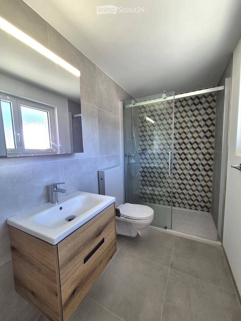 Modern bathroom with a white sink, wooden cabinet, toilet, glass-enclosed shower, and a window providing natural light.