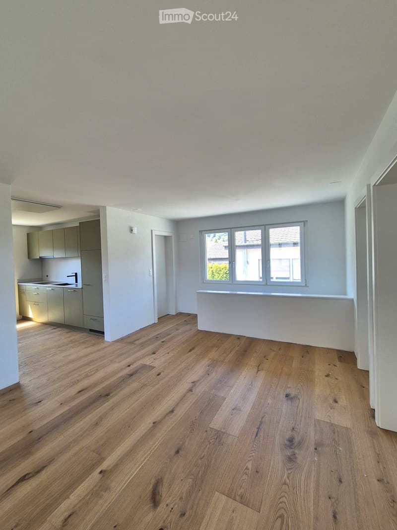 Empty room with light-colored wooden flooring, white walls, and a large window with a view of a neighboring roof. A small kitchen area is visible in the background with cabinets and a sink.