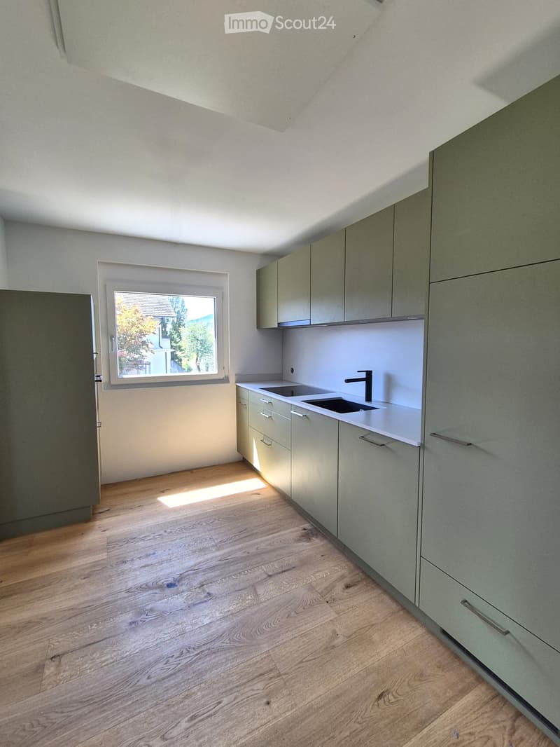 Modern kitchen with light grey cabinets, white countertops, and a double sink. The room features wooden flooring and a window letting in natural light.
