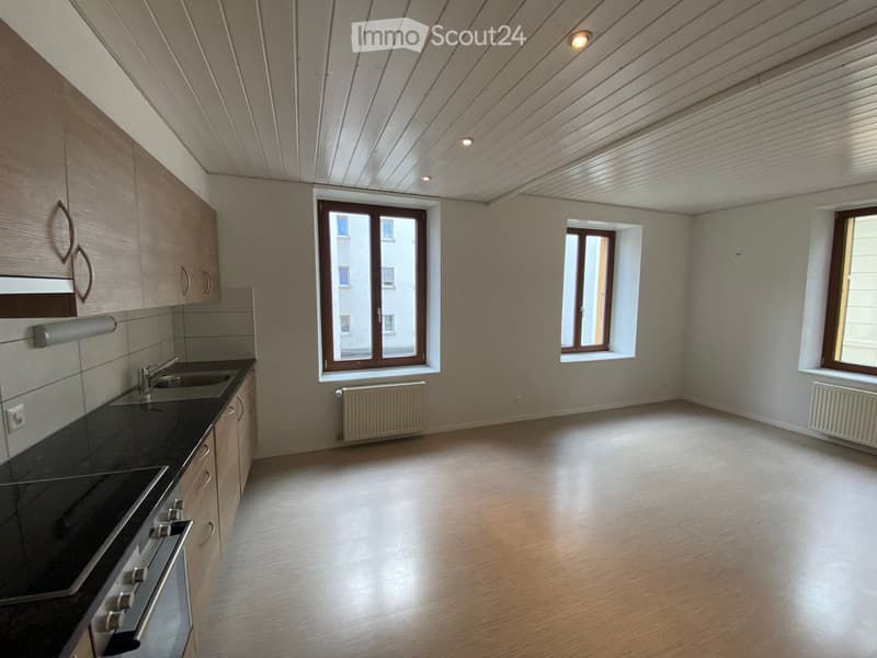 Empty kitchen with wooden cabinets, black countertop, sink, and three windows, featuring a light wooden floor and white walls.
