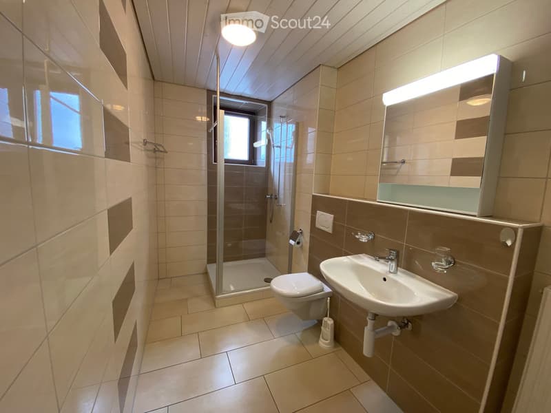 Modern bathroom with beige tiles, a toilet, a sink with a mirror, a glass-enclosed shower, and a window providing natural light.