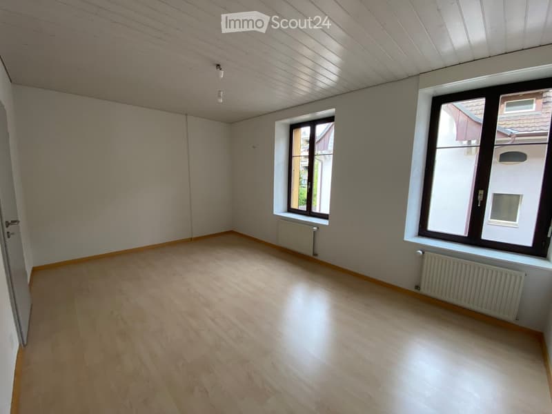 Empty room with wooden flooring, white walls, and large windows with black frames. Radiators are visible under the windows.