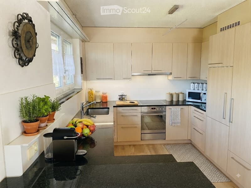 Modern kitchen with light wood cabinets, stainless steel appliances, black granite countertop, and various kitchen items including a coffee maker, fruit bowl, and potted plants.