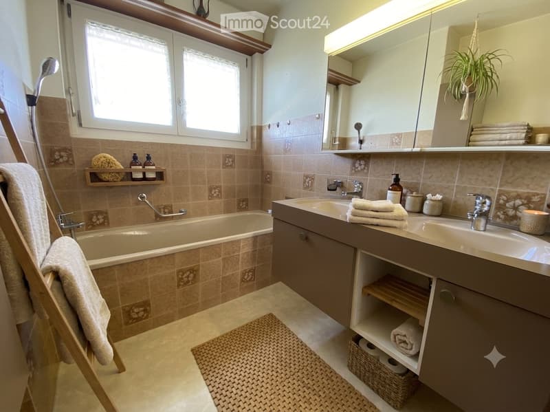 Modern bathroom with a bathtub, double sink, large mirror, and wooden shelves. Features include a shower, towels, and decorative items.