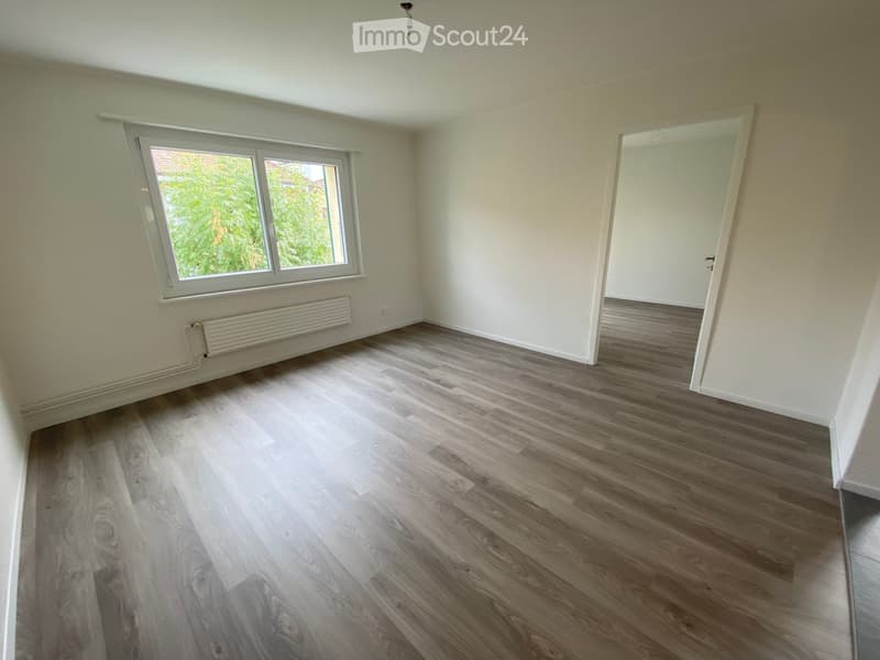 Empty room with wooden flooring, white walls, a window with white frame, and a radiator below the window. There is a doorway leading to another room.
