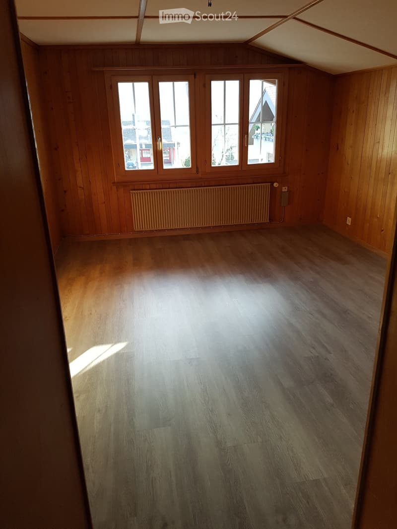 Empty room with wooden walls and flooring, large windows, and a radiator. No furniture or equipment present.