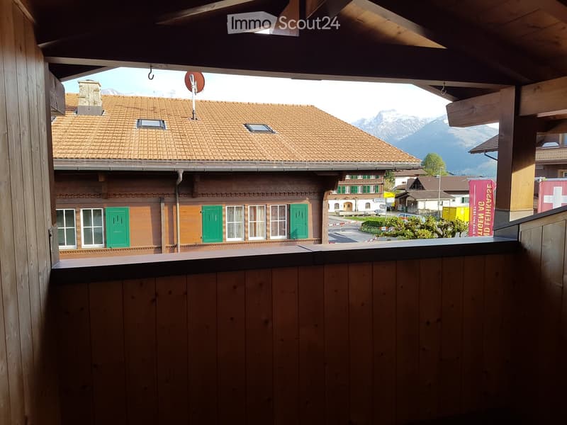 Balcony with wooden railing, overlooking a village with traditional houses, green shutters, and distant mountains.