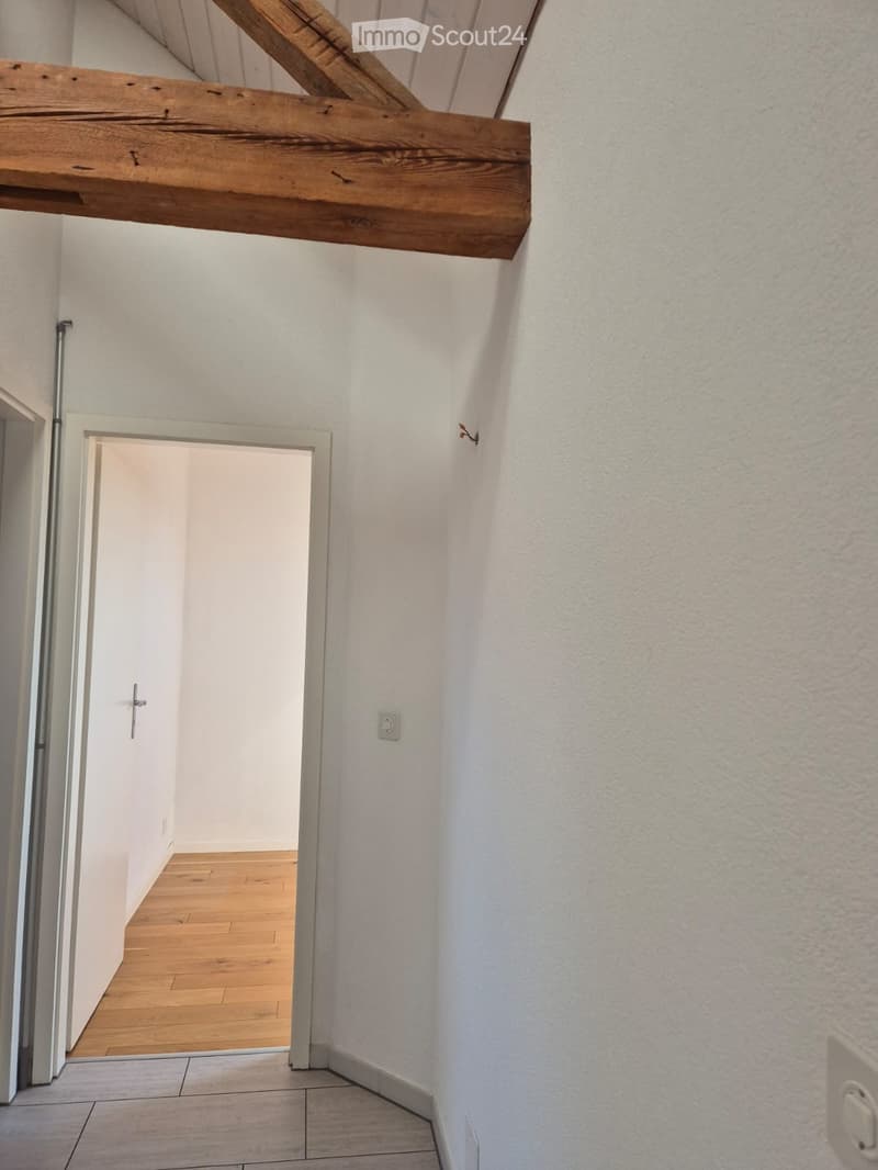 Empty hallway with white walls, wooden floor, and a wooden beam on the ceiling. A doorway leads to another room with wooden flooring.