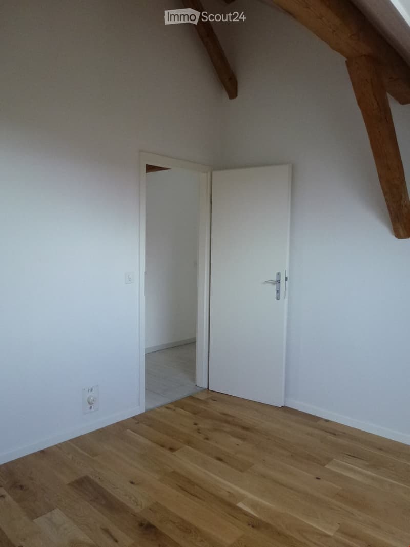 Empty attic room with wooden flooring, white walls, and exposed wooden beams. Features a closed white door and an open doorway leading to another space.