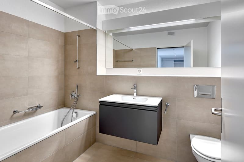 Modern bathroom with beige tiles, a bathtub, a sink with a dark cabinet, a toilet, and a large mirror with built-in lighting.