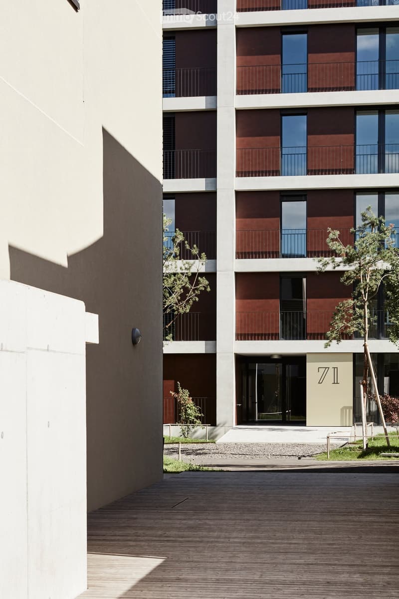Modern multi-story residential building with red and white facade, balconies, and small trees in the foreground.