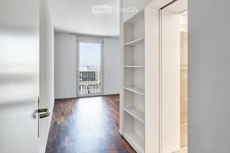 Empty room with wooden flooring, white walls, and a sliding glass door leading to a balcony with a view of the city.