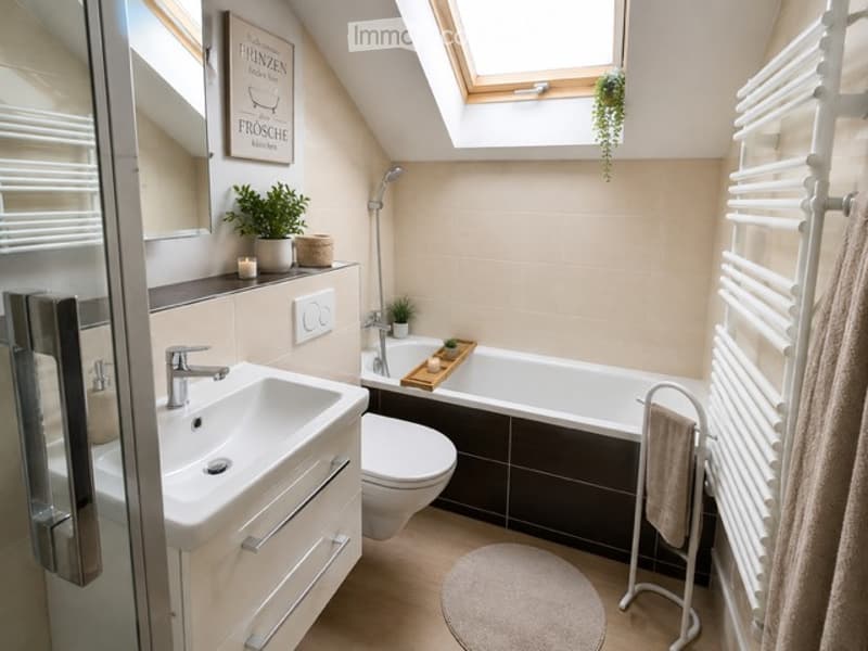 Modern bathroom with a white sink, toilet, bathtub, and towel rack. Features a skylight window, potted plants, and a decorative candle on the shelf.