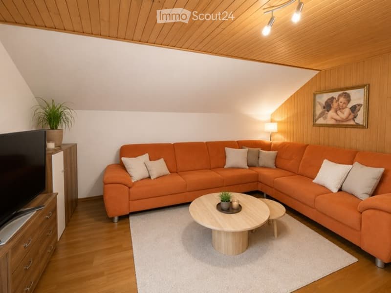 Modern living room with wooden ceiling and flooring, orange sectional sofa, white throw pillows, round wooden coffee table, side table, floor lamp, potted plant, and picture frame on the wall.