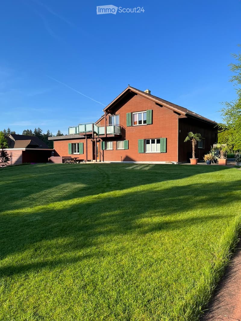 Two-story red brick house with green shutters, large windows, and a balcony on the upper level. The house is surrounded by a well-maintained lawn and features potted plants near the entrance.